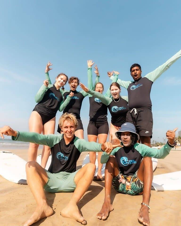 A beginner surf group in The Surfer rash guards celebrating on the sand at Weligama, Sri Lanka