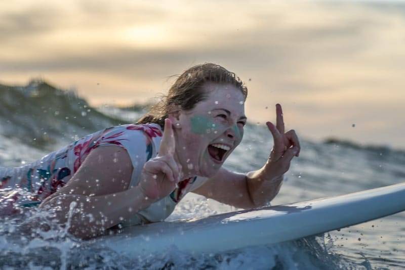 Eine Anfängerin liegt lachend auf ihrem Surfboard bei Sonnenuntergang während eines Kurses in Weligama