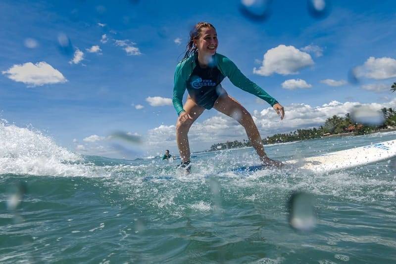 Eine Surferin im türkisfarbenen Lycra steht auf einer Anfängerwelle in der Weligama Bay auf