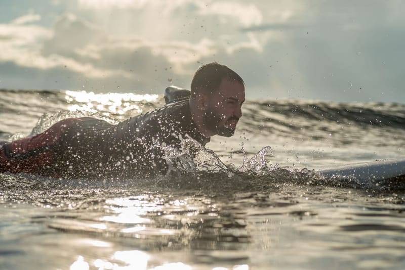 Ein Surfer paddelt bei Gegenlicht durch die Gischt in Weligama