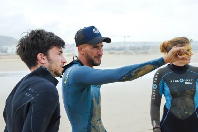 Ein Surfcoach erklärt zwei Schülern die Bedingungen am Strand von Tamraght, Marokko